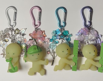 May include: Four keychains featuring pale yellow figures attached to silver, pink, blue, and purple carabiners. The figures hold miniature green tools including a shovel, a hard hat, and rulers. Each keychain is adorned with colorful beads and charms.