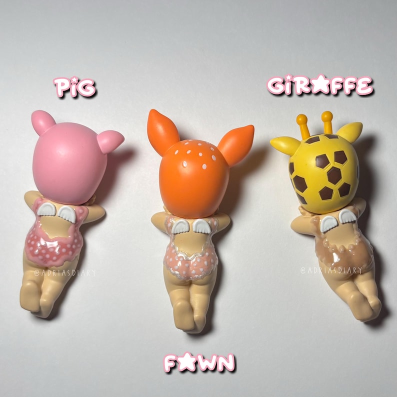 Puede incluir: Tres peque&ntilde;as figuras: un cerdo rosa, un cervatillo naranja y una jirafa amarilla. Cada figura tiene un bikini y posa con los brazos cruzados. La palabra "PIG" est&aacute; encima del cerdo rosa, "GIRAFFE" encima de la jirafa y "FAWN" debajo del cervatillo naranja.