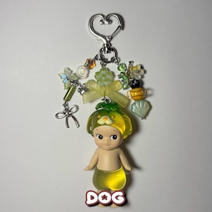 Puede incluir: Un llavero decorativo con una peque&ntilde;a figura de perro con sombrero y falda amarillos. El llavero incluye un cierre en forma de coraz&oacute;n, cuentas y abalorios de varias formas y colores, incluyendo una abeja y un lazo.