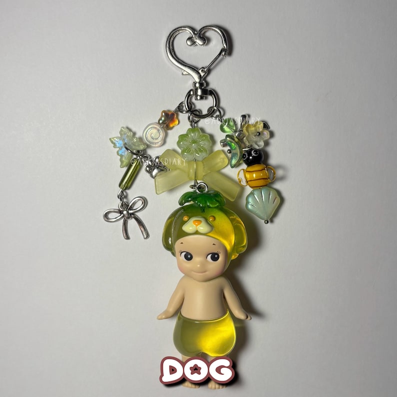 Puede incluir: Un llavero decorativo con una peque&ntilde;a figura de perro con sombrero y falda amarillos. El llavero incluye un cierre en forma de coraz&oacute;n, cuentas y abalorios de varias formas y colores, incluyendo una abeja y un lazo.