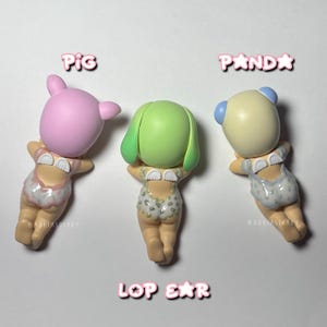 Puede incluir: Tres peque&ntilde;as figuras de animales: un cerdo rosa, un animal verde con orejas ca&iacute;das y un panda. Cada figura lleva un traje de ba&ntilde;o estampado. Las palabras "PIG", "PANDA" y "LOP EAR" est&aacute;n escritas encima y debajo de las figuras.