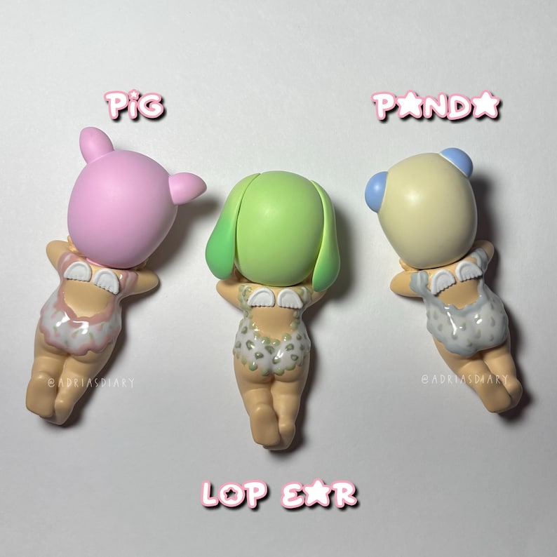 Puede incluir: Tres peque&ntilde;as figuras de animales: un cerdo rosa, un animal verde con orejas ca&iacute;das y un panda. Cada figura lleva un traje de ba&ntilde;o estampado. Las palabras "PIG", "PANDA" y "LOP EAR" est&aacute;n escritas encima y debajo de las figuras.