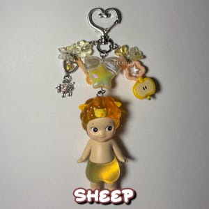 Puede incluir: Un llavero con una peque&ntilde;a figura de beb&eacute; con una melena de le&oacute;n naranja y una falda amarilla. El llavero incluye un cierre en forma de coraz&oacute;n, un dije de oveja y varios dijes decorativos como una estrella y una manzana. La palabra "SHEEP" est&aacute; escrita en la parte inferior.