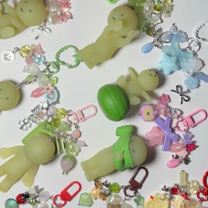 May include: A collection of whimsical keychains featuring small, light green figures in various poses. Each keychain includes colorful beads, charms, and a lobster clasp. Some figures wear green hats or hold green objects. The keychains have pink, red, and brown clasps.