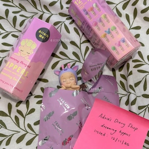 K&ouml;nnte beinhalten: Eine Sammlung von Sonny Angel HIPPERS-Figuren, darunter eine kleine, schlafende Figur mit einem lila-blauen Giraffenhut. Das Bild zeigt rosa Schachteln und Verpackungen mit dem Text "Sonny Angel HIPPERS" und "Dreaming Series".