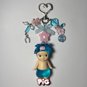 Puede incluir: Un llavero decorativo con una peque&ntilde;a figura que lleva un sombrero y pantalones cortos de cerdo azules. El llavero tiene un cierre plateado en forma de coraz&oacute;n y est&aacute; adornado con cuentas azules, rosas y transparentes, un lazo y una estrella. La palabra "PIG" est&aacute; en la parte inferior.