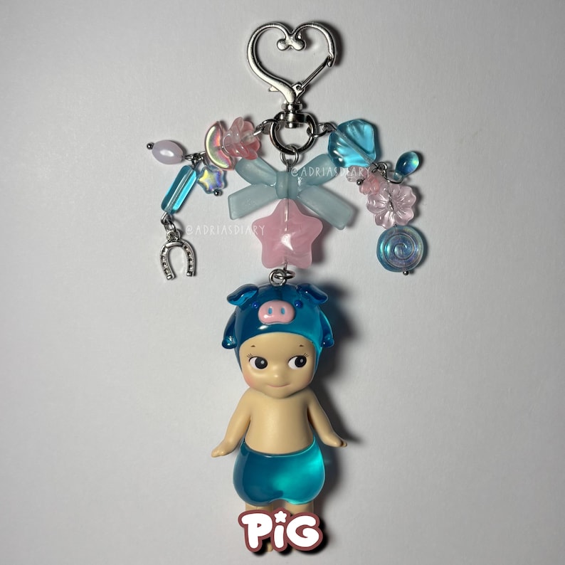 Puede incluir: Un llavero decorativo con una peque&ntilde;a figura que lleva un sombrero y pantalones cortos de cerdo azules. El llavero tiene un cierre plateado en forma de coraz&oacute;n y est&aacute; adornado con cuentas azules, rosas y transparentes, un lazo y una estrella. La palabra "PIG" est&aacute; en la parte inferior.