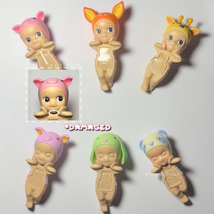 Puede incluir: Una colecci&oacute;n de peque&ntilde;as figuras de color canela, cada una adornada con un sombrero con tem&aacute;tica de animales. Los sombreros incluyen un cerdo, un ciervo, una jirafa, un panda y un conejo. La palabra "DAMAGED" tambi&eacute;n es visible.