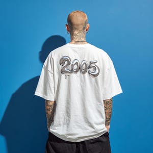 Puede incluir: Camiseta blanca con el número "2005" plateado metálico impreso en la espalda. La camiseta tiene un corte holgado y mangas cortas. El fondo es azul liso.