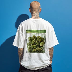 May include: White t-shirt with a graphic of a clear plastic bag filled with green cannabis buds. The bag has a green zip closure. The t-shirt is oversized with short sleeves. The background is a solid blue.