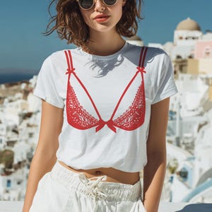 以下が含まれることがあります： 赤いブラジャーのデザインが施された白いTシャツ。ブラジャーのグラフィックには、レースのディテールとリボンが含まれています。半袖のTシャツは、サングラスをかけている人が着用しています。
