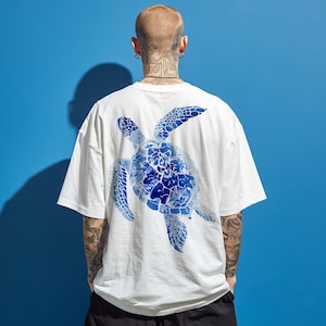 May include: White t-shirt with a large blue sea turtle design on the back. The turtle is filled with a floral pattern. The shirt has short sleeves and a relaxed fit. The background is a solid blue.