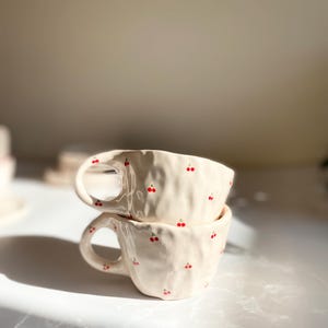 Może przedstawiać: Dwa białe ceramiczne kubki z czerwonym wzorem wiśni ułożone jeden na drugim. Kubki mają fakturowaną powierzchnię.
