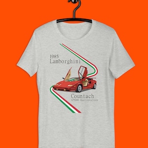 Puede incluir: Camiseta gris claro con un Lamborghini Countach rojo. El diseño incluye el texto "1985 Lamborghini Countach LP5000 Quattrovalvole" y una franja verde, blanca y roja.