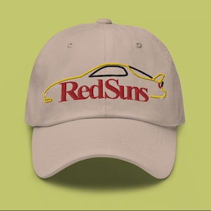 Puede incluir: Una gorra de béisbol beige con una silueta de coche amarilla bordada y el texto "RedSuns" en rojo.