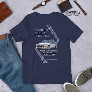 Puede incluir: Una camiseta azul marino con un gráfico de coche blanco y el texto "Lancia Delta Integrale" y "Most Successful Rally Car of All Time". El texto "FastLaneFashionAuto" está en la parte inferior de la camiseta.