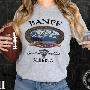 May include: A light gray long-sleeve sweatshirt with the word "BANFF" above an oval graphic of a mountain scene. Below the graphic are the words "Outdoor Discovery", "Canadian Tradition", and "ALBERTA". A brown football is held in the left hand.