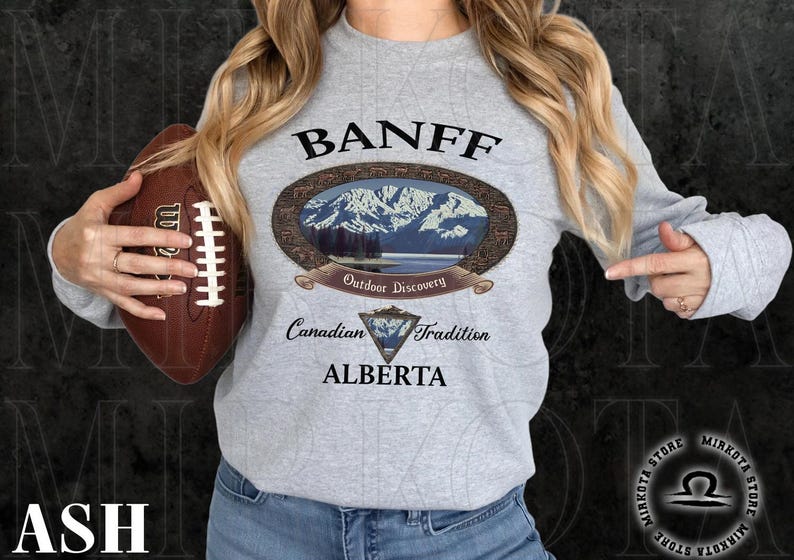 May include: A light gray long-sleeve sweatshirt with the word "BANFF" above an oval graphic of a mountain scene. Below the graphic are the words "Outdoor Discovery", "Canadian Tradition", and "ALBERTA". A brown football is held in the left hand.