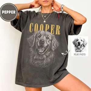 Op de afbeelding: Donkergrijs t-shirt met een gedetailleerde illustratie van een Golden Retriever en de naam "COOPER" in gouden letters. Een kleine afbeelding van een hond met de tekst "YOUR PHOTO" staat rechts.