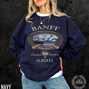 May include: Navy blue sweatshirt with a white collar featuring a Banff, Alberta design. The design includes an oval graphic with a mountain and lake scene, the words "Outdoor Discovery" and "Canadian Tradition."