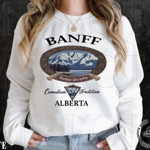 May include: White sweatshirt featuring a Banff, Alberta design. The design includes an oval image of mountains and a lake, with text that reads "Outdoor Discovery" and "Canadian Tradition". Below the oval is a diamond-shaped emblem and the word "ALBERTA".