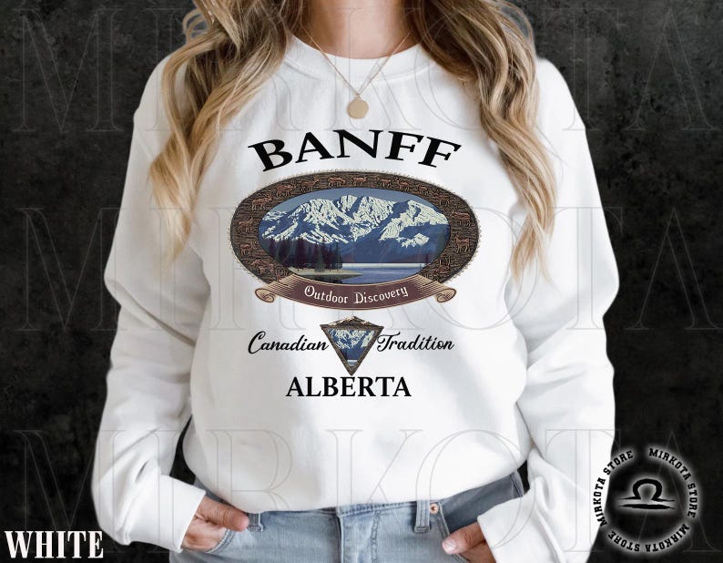 May include: White sweatshirt featuring a Banff, Alberta design. The design includes an oval image of mountains and a lake, with text that reads "Outdoor Discovery" and "Canadian Tradition". Below the oval is a diamond-shaped emblem and the word "ALBERTA".