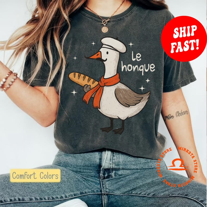 K&ouml;nnte beinhalten: Ein dunkelgraues T-Shirt mit einer Cartoon-Gans, die einen Kochhut tr&auml;gt und eine Baguette h&auml;lt. Der Text "le honque" ist auf dem Shirt aufgedruckt.