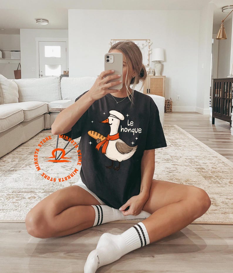 K&ouml;nnte beinhalten: Ein schwarzes T-Shirt mit einem Cartoon-Gans, die einen Kochhut tr&auml;gt und eine Baguette h&auml;lt. Der Text "Le honque" ist auf dem Shirt gedruckt.