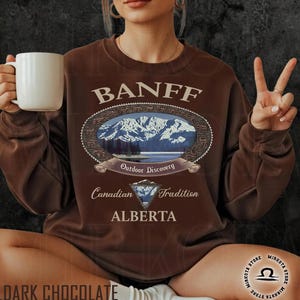 May include: Dark chocolate brown sweatshirt with the word "BANFF" above an oval graphic of a mountain scene. The graphic includes the words "Outdoor Discovery", "Canadian Tradition", and "ALBERTA". A white mug is held in the left hand.