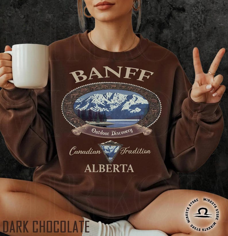 May include: Dark chocolate brown sweatshirt with the word "BANFF" above an oval graphic of a mountain scene. The graphic includes the words "Outdoor Discovery", "Canadian Tradition", and "ALBERTA". A white mug is held in the left hand.