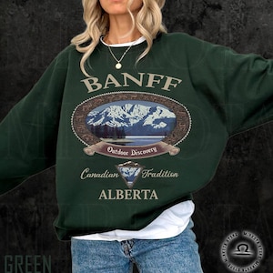 May include: A dark green sweatshirt with the word "BANFF" in a light brown font. The sweatshirt features an oval graphic with a mountain landscape and the words "Outdoor Discovery", "Canadian Tradition", and "ALBERTA".