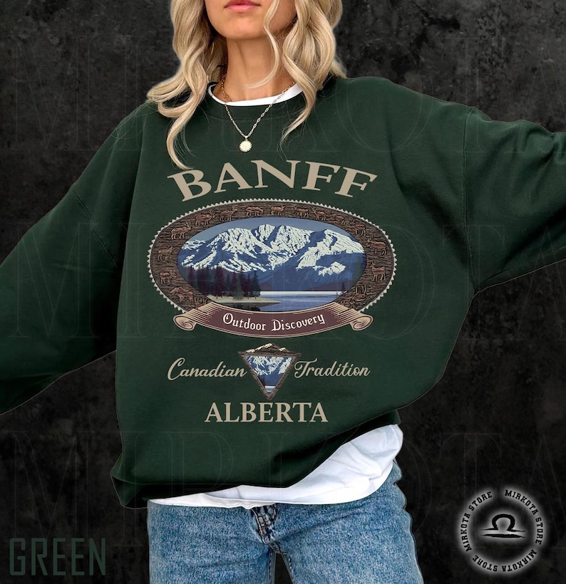May include: A dark green sweatshirt with the word "BANFF" in a light brown font. The sweatshirt features an oval graphic with a mountain landscape and the words "Outdoor Discovery", "Canadian Tradition", and "ALBERTA".