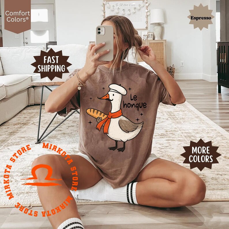 K&ouml;nnte beinhalten: Ein braunes T-Shirt mit einer Cartoon-Illustration einer Gans, die einen Kochhut tr&auml;gt und eine Baguette h&auml;lt. Der Text "Le honque" ist auf dem Shirt aufgedruckt.