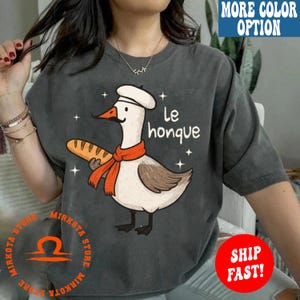 K&ouml;nnte beinhalten: Ein graues T-Shirt mit einem Cartoon-Gans, die einen Kochhut tr&auml;gt und eine Baguette h&auml;lt. Der Text "Le honque" ist auf dem Shirt gedruckt.