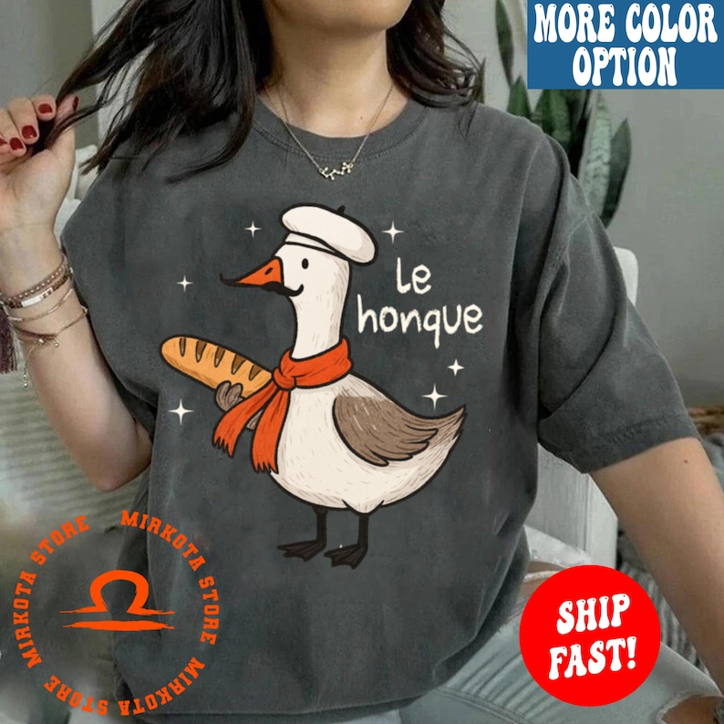 K&ouml;nnte beinhalten: Ein graues T-Shirt mit einem Cartoon-Gans, die einen Kochhut tr&auml;gt und eine Baguette h&auml;lt. Der Text "Le honque" ist auf dem Shirt gedruckt.