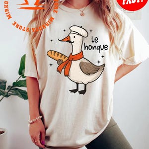 K&ouml;nnte beinhalten: Ein wei&szlig;es T-Shirt mit einer Cartoon-Illustration einer Gans, die einen Kochhut tr&auml;gt und eine Baguette h&auml;lt. Der Text "Le honque" ist auf das Shirt gedruckt.