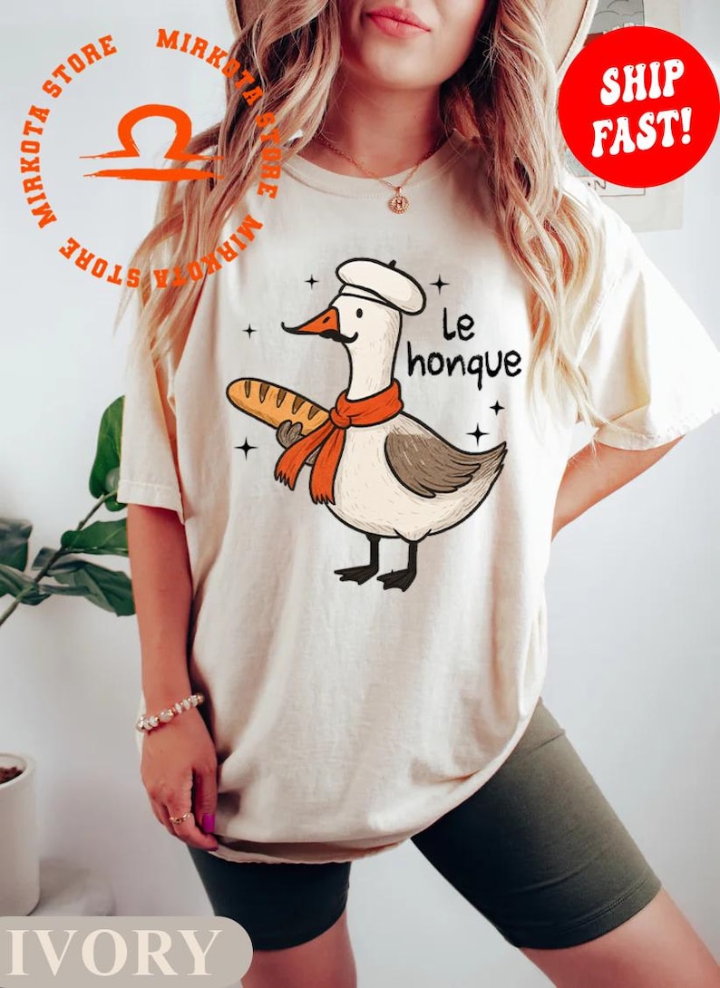 K&ouml;nnte beinhalten: Ein wei&szlig;es T-Shirt mit einer Cartoon-Illustration einer Gans, die einen Kochhut tr&auml;gt und eine Baguette h&auml;lt. Der Text "Le honque" ist auf das Shirt gedruckt.