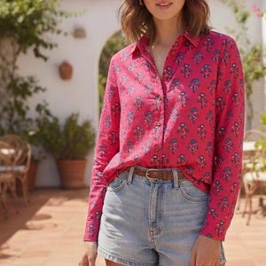 Op de afbeelding: Een overhemd met lange mouwen en knoopsluiting in een levendige roze kleur met een herhalend patroon van kleine blauwe bloemen. Het overhemd wordt gecombineerd met een lichtblauwe spijkerbroek en een dunne bruine gevlochten leren riem met een goudkleurige gesp.