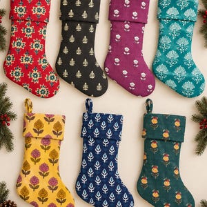 Könnte beinhalten: Sieben Weihnachtsstrümpfe in zwei Reihen angeordnet, jeder mit einem einzigartigen Blockdruckmuster. Die Farben umfassen Rot, Schwarz, Lila, Türkis, Gelb, Marineblau und Dunkelgrün, jeweils mit einem umgeschlagenen Bündchen und einer Aufhängeschlaufe.