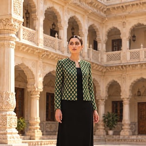 May include: A long-sleeved, open-front jacket in dark green with a repeating gold-toned teardrop pattern, worn over a simple, floor-length black dress. The jacket features a round neckline and a structured, straight-cut silhouette.