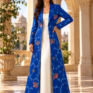 May include: A floor-length royal blue coat featuring a vibrant floral pattern with pink and orange accents, worn over a simple white sleeveless dress. The coat has long sleeves, a structured collar, and a straight, open-front silhouette.