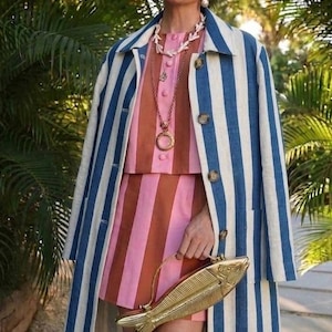 May include: A woman wearing a striped blue and white coat over a pink and brown striped dress. She is holding a gold fish-shaped handbag and wearing jewelry. The outfit is set against a backdrop of green foliage.