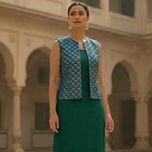 May include: A woman wearing a sleeveless teal vest with a gold geometric pattern over a long, emerald green dress. The outfit is set against a backdrop of a building with arched doorways.
