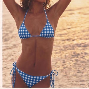 May include: A two-piece bikini set featuring a blue and white gingham check pattern. The top is a triangle style with thin halter straps, and the bottom is a classic cut with adjustable side ties that are knotted into bows on each hip.