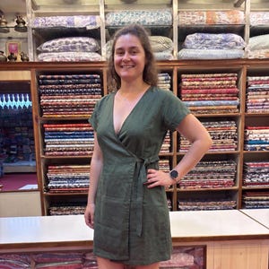 Könnte beinhalten: Ein olivgrünes Wickelkleid mit kurzen Ärmeln, V-Ausschnitt und Bindeband. Das Kleid ist aus einem strukturierten Stoff gefertigt. Die Person steht vor Regalen mit bunten Textilien. Eine Uhr ist am Handgelenk zu sehen.