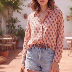 May include: A long-sleeved button-down shirt in a peach-coloured fabric featuring a repeating dark blue and white floral pattern, paired with light blue denim shorts and a thin brown braided leather belt with a silver-toned metal buckle.