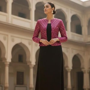 May include: A woman's outfit featuring a black maxi dress paired with a magenta jacket. The jacket has a detailed pattern and long sleeves. The background shows an architectural structure with arches and columns.