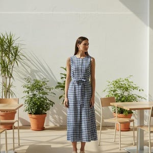 Könnte beinhalten: Ein ärmelloses, blau-weiß kariertes Kleid mit passendem Gürtel. Das Kleid ist knielang und wird mit neutralen Sandalen getragen. Die Umgebung ist im Freien, mit Topfpflanzen und Tischen und Stühlen aus Holz.