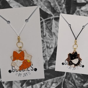 May include: Two pendant necklaces with animal designs. One pendant is an orange, white, and yellow fox, and the other is a black and white cat. Both pendants are on white cards with the text "Goodies by Jay."