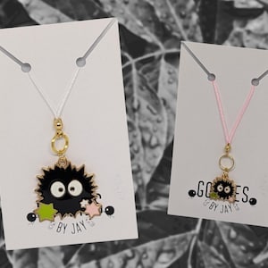 May include: Two necklaces with black and green charms on white and pink card stock backgrounds. The charms are of a black furry creature with large eyes and green stars. The text on the cards reads "Goodies by Jay."
